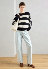 Pulôver de malha riscada em preto e creme, jeans de perna larga azul claro e botas de tornozelo de salto castanhas. Modelo segura uma mala de ombro creme.