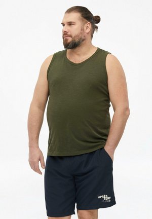 Brodaty mężczyzna z włosami związanymi w koczek, ubrany w oliwkowozieloną bezrękawnik i granatowe krótkie spodenki, stoi z jedną ręką w kieszeni.