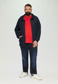 Navy zip-up hoodie over een rood t-shirt, gecombineerd met donkere jeans en witte sneakers. De hoodie heeft een voorzak en subtiele accenten.