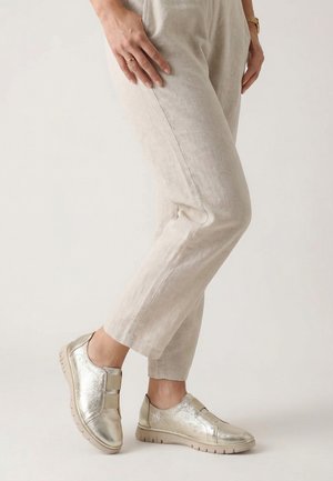 Person, die beige Hose und metallische Slipper trägt, steht mit einem Fuß leicht erhoben vor einem einfarbigen hellen Hintergrund.