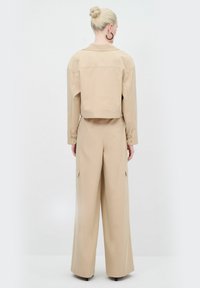 Giacca corta beige abbinata a pantaloni palazzo a vita alta. Entrambi realizzati in tessuto liscio, con tasche a toppa e dettagli con bottoni.