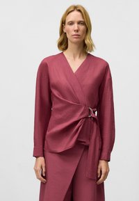 Hallonfärgad linnes wrap-topp med långa ärmar och en frontknut med en silverring som accent. Slät textur och minimalistisk design.