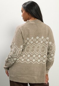 Beige gebreide trui met een wit geometrisch en bloemmotief, geribbelde manchetten en een hoge halslijn, met een relaxte pasvorm aan de achterkant.