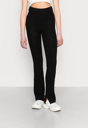 Leggings pretos de malha com um padrão texturizado e rachas laterais na barra, combinados com ténis brancos. Cintura elástica para maior conforto.