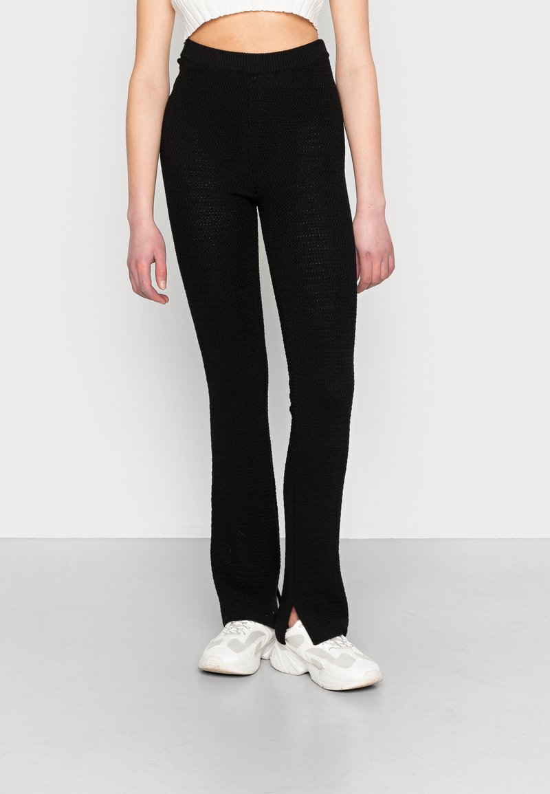 Leggings pretos de malha com um padrão texturizado e rachas laterais na barra, combinados com ténis brancos. Cintura elástica para maior conforto.