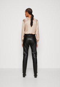 Blouse en satin beige avec détail froncé au dos, associée à un pantalon en cuir noir taille haute. Des bottes à talons sombres accentuent la tenue.