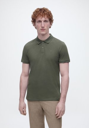 Młody mężczyzna z kręconymi rudymi włosami, ubrany w ciemnozieloną koszulkę polo i beżowe spodnie, stojący na tle jednolitego jasnego tła.