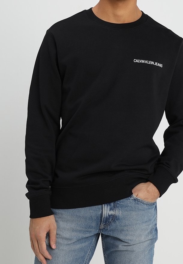 Mustavillainen puuvillapaita pitkähihaisilla hihoilla ja pyöreällä kaula-aukolla, jossa vasemmassa rinnassa on valkoinen "Calvin Klein Jeans" -logo.
