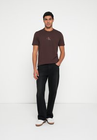 Hnědé tričko s kulatým výstřihem a malým logem CK, doplněné o černé rozšířené džíny a bílé tenisky, na jednoduchém pozadí.