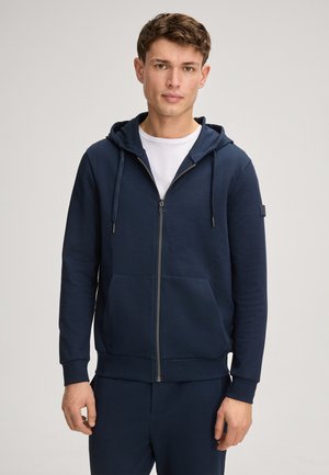 Hoodie zippé bleu marine en coton doux, avec une poche avant, une capuche à cordon de serrage et des poignets côtelés. Porté par-dessus une chemise blanche.