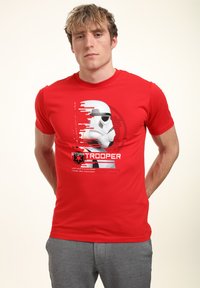 Czerwony t-shirt z krótkim rękawem, z grafiką hełmu szturmowca, z wyraźnymi czarnymi i szarymi akcentami, na białym tle.