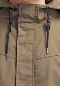 Olivově zelená bunda s vysokým límcem, která má černé stahovací šňůrky, plastové brzdičky a dvě náprsní kapsy s hladkým, strukturovaným povrchem.