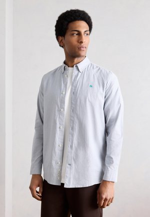 Man with afro hairstyle wearing light blue button-up shirt over white t-shirt looking to the side against plain background