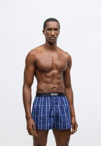 Modre karo boksar hlače s črnim pasom, označenim z napisom "BOSS." Izdelane iz mehke tkanine, z gumbnim odpiranjem in sproščenim krojem.