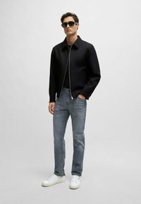 Giacca nera con zip sopra una maglietta nera, abbinata a jeans azzurri chiari e sneakers bianche. Occhiali da sole eleganti completano il look.