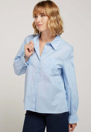 Chemise bleu clair à manches longues, avec des accents en dentelle texturée sur les poignets et la poche poitrine, fabriquée en tissu de coton lisse.