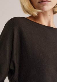 Sötétbarna pulóver, puha tapintással, széles, hajónyakú és esővállú kialakítással. Az anyag könnyűnek tűnik és enyhén textúrált.