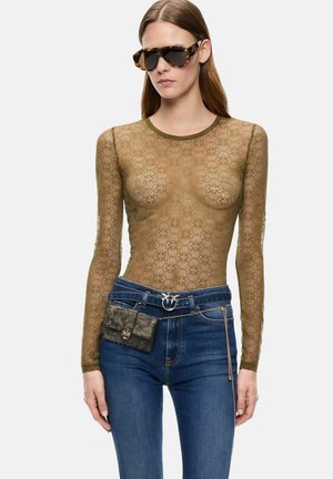 Donna che indossa occhiali da sole tartarugati, un top trasparente in pizzo oliva a maniche lunghe, jeans blu e una piccola borsa metallica da cintura con catena.