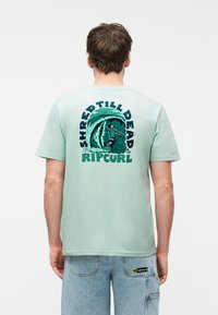 Νέος άνδρας που φοράει ανοιχτό πράσινο μπλουζάκι με γραφικό σχεδιασμό surfer και κύματος και κείμενο «Shred Till Dead Rip Curl» στην πλάτη, σε συνδυασμό με ανοιχτό τζιν σορτς.