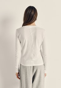 Žena s dugom smeđom kosom, odjevena u bijelu majicu s dugim rukavima i sive hlače, stoji okrenuta prema jednostavnoj svijetloj pozadini.