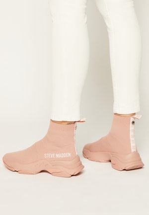 Baskets en tricot rose avec semelles épaisses portées avec un pantalon blanc ajusté, présentant la marque "Steve Madden" sur le côté extérieur et la languette arrière.