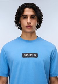 Ανοιχτό μπλε βαμβακερό μπλουζάκι με μαύρο ορθογώνιο λογότυπο και λευκό κείμενο "NAPA:PURI" στο μπροστινό μέρος, με ομαλή υφή και κανονική εφαρμογή.