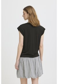 Ženska z ramenom dolgimi lasmi, oblečena v črno zgornji del brez rokavov in kratko plisirano karo krilo, obrnjena stran proti enostavni podlagi.