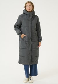 Manteau long gris rembourré avec un col montant, présentant un design matelassé, des poches latérales et des poignets côtelés, associé à un jean à jambes larges et des baskets.