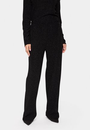 Pantalon noir à jambes larges, fabriqué dans un tissu texturé et légèrement brillant. Doté d'une taille élastique et d'une coupe décontractée, associé à des chaussures noires à bout pointu.