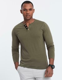 Man i en olivgrön långärmad henley tröja med tre knappar, vita byxor och svart armbandsur, stående med ena handen i fickan.