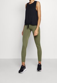 Zwarte mouwloze top met een geknoopt front, gecombineerd met olijfgroene leggings met diagonale stiksel. Zwarte sneakers met roze accenten.