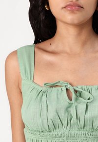 Haut vert clair en tissu texturé ; dispose de bretelles larges, d'un décolleté froncé et d'une petite ouverture en forme de clé à l'avant avec un détail de lien.