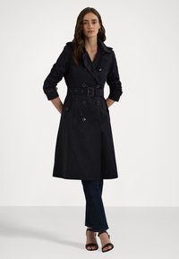 Zwarte trenchcoat met dubbele knopen, een ceintuur in de taille en een brede kraag. Gecombineerd met donkere jeans en sandalen met hakken.