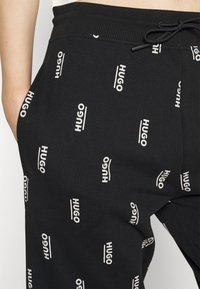 Zwarte joggers met een wit herhalend "HUGO" tekstpatroon. Voorzien van een elastische tailleband met trekkoord, zijzakken en een zachte stoftextuur.