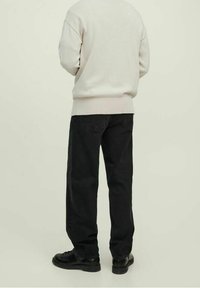 Person står med ryggen vänd, klädd i en off-white tröja, svarta raka jeans och svarta läderskor framför en ljus, enfärgad bakgrund.