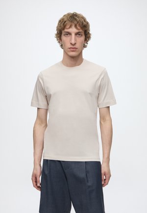 Ung mand med krøllet lyst brunt hår iført en ensfarvet beige t-shirt med korte ærmer og mørkegrå bukser, stående mod en hvid baggrund.
