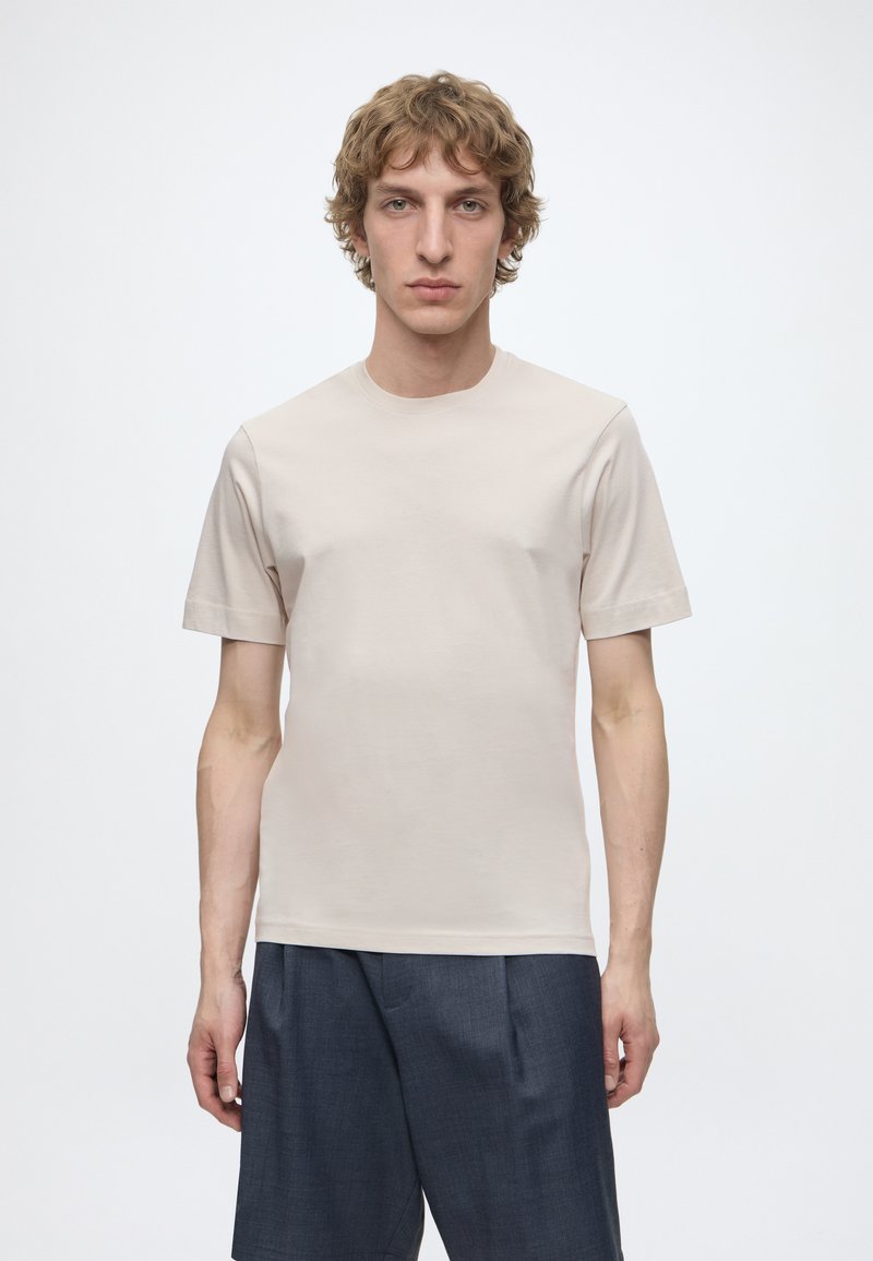 Jeune homme aux cheveux bouclés châtain clair portant un t-shirt beige uni à manches courtes et un pantalon gris foncé, debout devant un fond blanc.