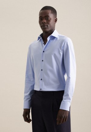 Camicia azzurro chiaro con maniche lunghe, caratterizzata da bottoni scuri a contrasto e dettagli sul colletto. Indossata con pantaloni scuri.