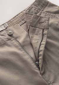 Pantalones beige con cremallera abierta, botón y cierre de velcro que muestran costuras detalladas y textura de la tela.