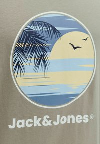 Coucher de soleil sur l'océan avec des feuilles de palmier et deux oiseaux, encadré dans un cercle au-dessus du texte "Jack & Jones" sur un fond en tissu.