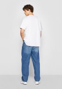 Hvit bomull T-skjorte med korte ermer og avslappet passform, kombinert med løse lys blå denim jeans og hvite sneakers. Modellen står med ryggen til.
