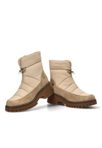 Botines beige con un diseño acolchado, acentos de ante y una suela de goma texturizada. Presenta un cierre con cremallera y herrajes decorativos.