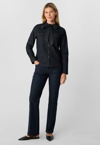 Donkerdenim overhemd met een strikjeskraag, twee borstzakken en knoopsluiting. Gecombineerd met een donkerdenim flared broek en zwarte slingback schoenen.
