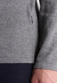 Pull-over en maille grise avec finition texturée, coutures latérales détaillées et une petite étiquette de marque sur le côté gauche. Tissu lisse et coupe ajustée.