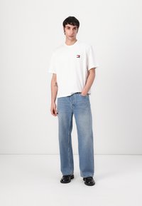 Hvid T-shirt med korte ærmer og et lille logo på brystet, parret med lysblå lige ben-jeans og sorte sko. Simpelt design.