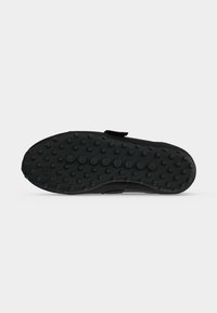 Semelle d'une chaussure noire avec des grips ronds espacés uniformément, présentée sur un fond clair uni.