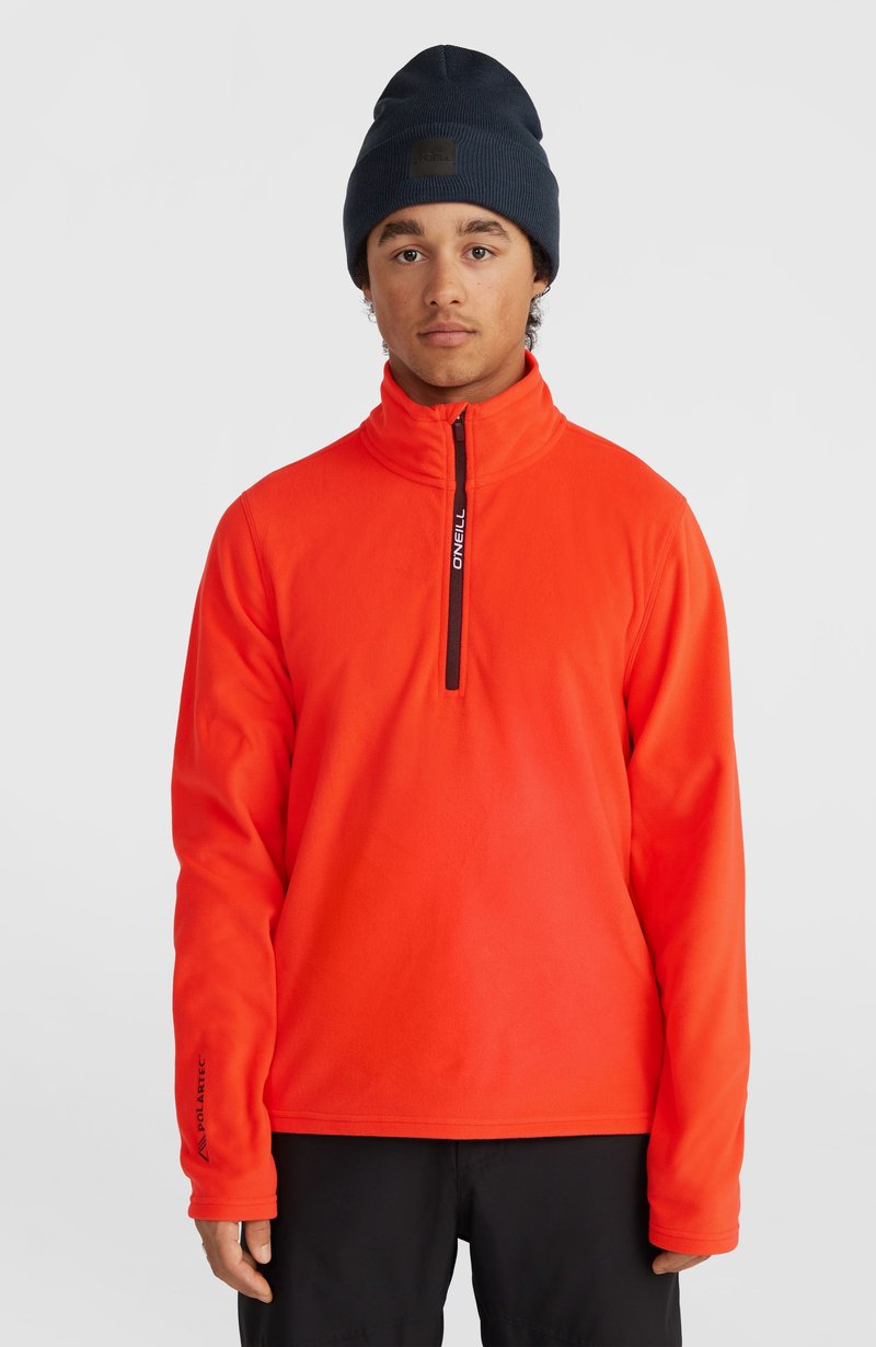 Pomarańczowy polarowy sweter z wysokim kołnierzem, stylu ćwierć-zamek, z czarnym zamkiem błyskawicznym i logo, zestawiony z granatowym beanie.