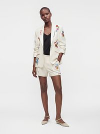 Femme portant un blazer blanc brodé et un short assorti avec un haut noir et des ballerines beige à bout pointu, debout avec une main dans la poche.