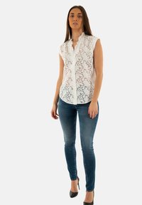Witte mouwloze blouse met geborduurde bloemen-uitsparingen, ruches bij de kraag, gecombineerd met strakke blauwe jeans en zwarte hoge hakken.