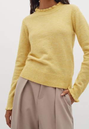 Femme portant un pull doux à manches longues jaune pâle associé à un pantalon beige taille haute, une main dans la poche et l'autre détendue.